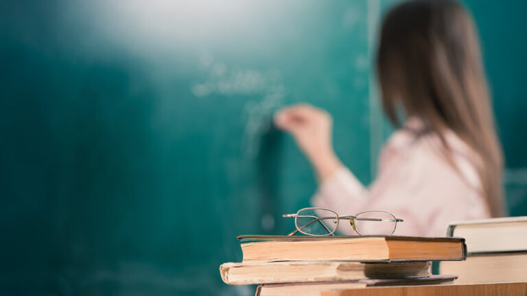 Teacher writing on a classroom board with books and glasses in the foreground.