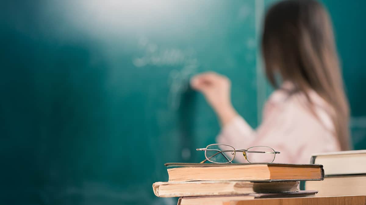Teacher writing on a classroom board with books and glasses in the foreground.